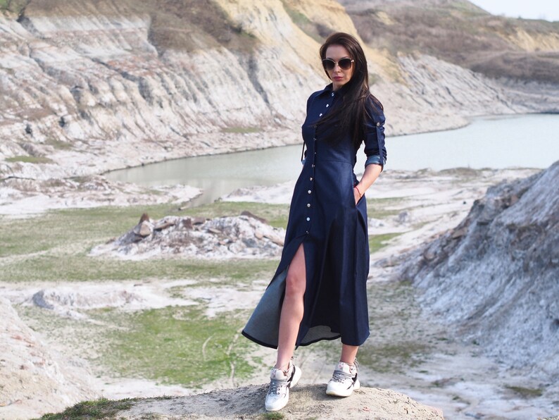 May include: A woman wearing a dark blue denim dress with a long slit up the side and white sneakers. She is standing on a rocky hill with a body of water in the background.