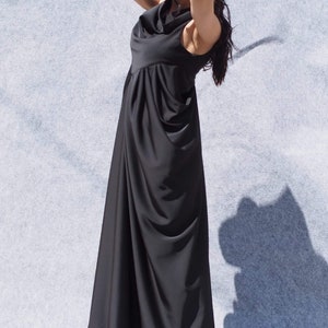 May include: A woman wearing a long black maxi dress with a cowl neck and a gathered skirt. She is wearing sunglasses and has her hair pulled back.