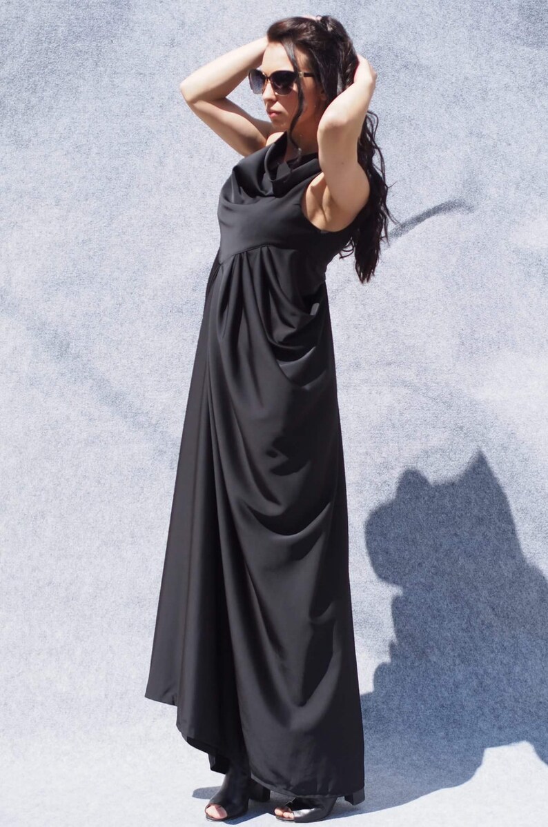 May include: A woman wearing a long black maxi dress with a cowl neck and a gathered skirt. She is wearing sunglasses and has her hair pulled back.