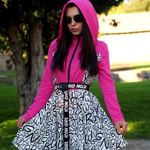 May include: A pink hooded jacket with a black zipper and a white and black skirt with a black belt that says "ICON GIRL" repeated. The skirt has a pattern of black letters on a white background.