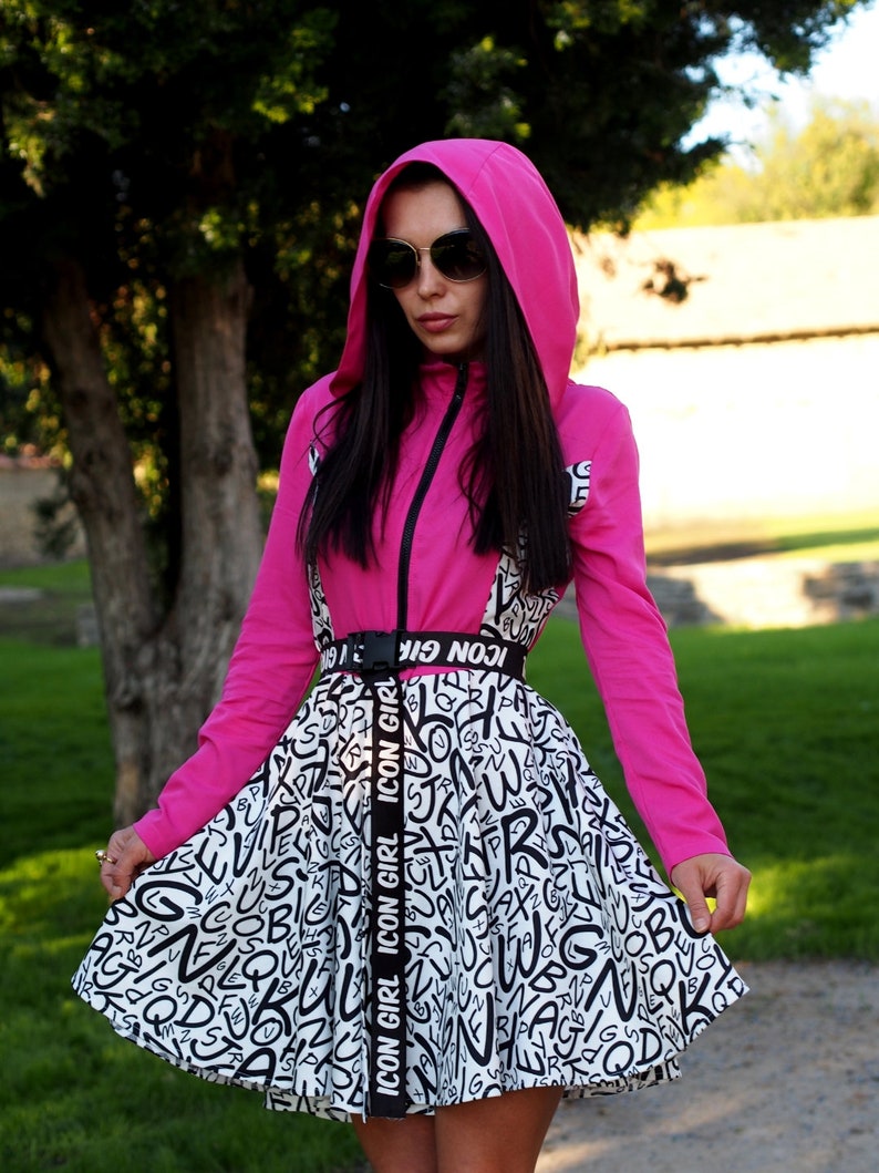 May include: A pink hooded jacket with a black zipper and a white and black skirt with a black belt that says "ICON GIRL" repeated. The skirt has a pattern of black letters on a white background.