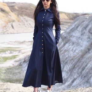 May include: A long, dark blue denim dress with a button-up front and long sleeves. The dress has a fitted bodice and a flowing skirt. The model is wearing white sneakers.