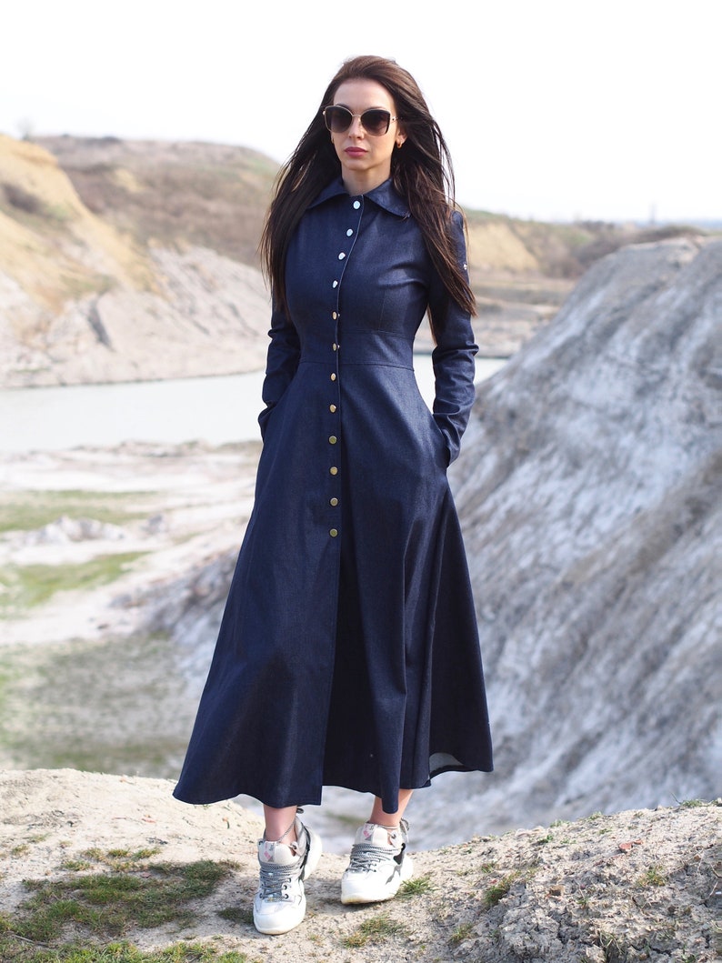 May include: A long, dark blue denim dress with a button-up front and long sleeves. The dress has a fitted bodice and a flowing skirt. The model is wearing white sneakers.