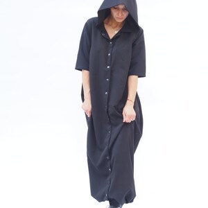Puede incluir: Un vestido negro con capucha, botones en la parte delantera y mangas largas. El vestido tiene un corte holgado y una silueta fluida.