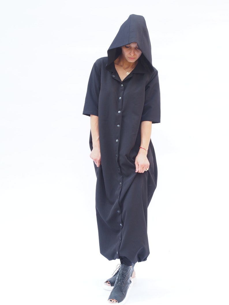 Puede incluir: Un vestido negro con capucha, botones en la parte delantera y mangas largas. El vestido tiene un corte holgado y una silueta fluida.