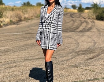 May include: A black and white houndstooth blazer dress with long sleeves. The dress is paired with black knee-high boots and a black beret. The background is a dirt road under a blue sky.