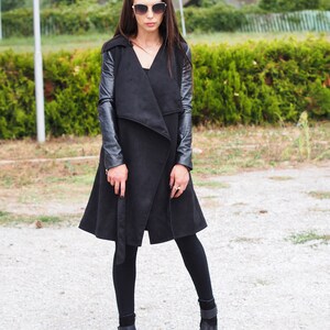 May include: A black long-sleeved coat with leather sleeves. The coat has a wrap-style closure and a belt. The model is wearing black leggings and black ankle boots.