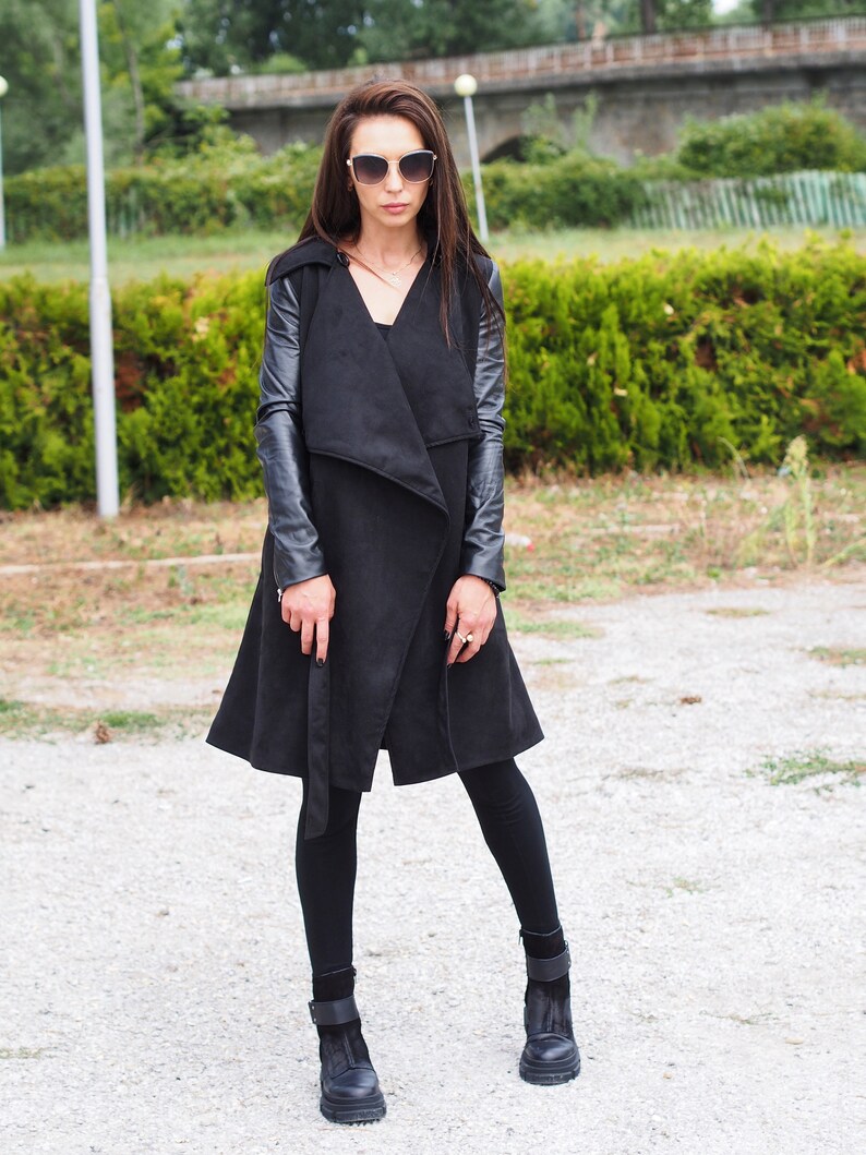 May include: A black long-sleeved coat with leather sleeves. The coat has a wrap-style closure and a belt. The model is wearing black leggings and black ankle boots.