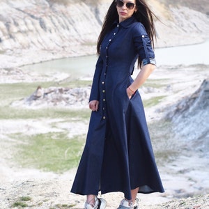 May include: A woman wearing a long, dark blue denim dress with a button-down front and long sleeves. She is wearing white sneakers and sunglasses.