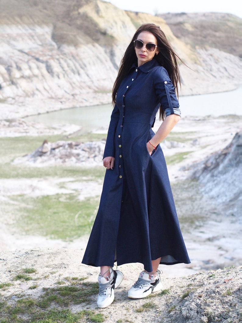 May include: A woman wearing a long, dark blue denim dress with a button-down front and long sleeves. She is wearing white sneakers and sunglasses.