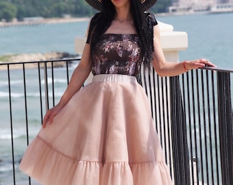 May include: A woman wearing a brown floral print top with black straps and a wide-brimmed straw hat with a black ribbon. She is wearing a light pink tulle skirt with a ruffled hem. The skirt is knee-length and has a fitted waistband.