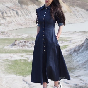 May include: A long, dark blue denim dress with a button-up front and a slightly flared skirt. The dress has long sleeves and a collared neckline. The model is wearing white sneakers with floral accents.