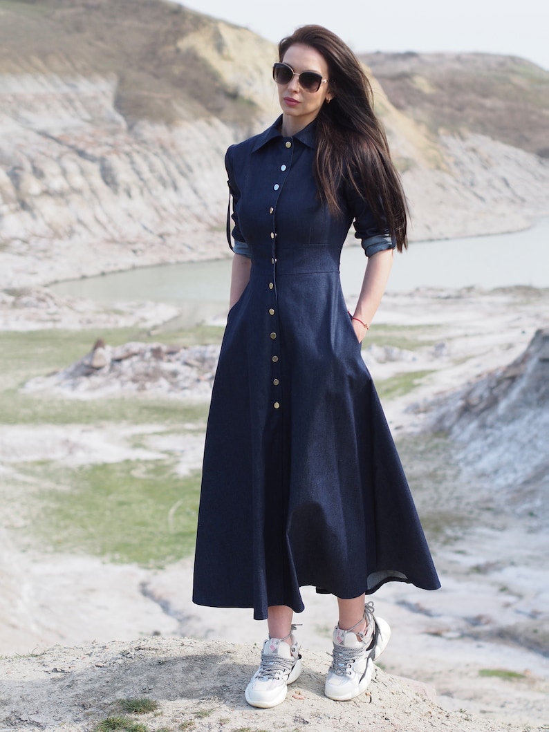 May include: A long, dark blue denim dress with a button-up front and a slightly flared skirt. The dress has long sleeves and a collared neckline. The model is wearing white sneakers with floral accents.