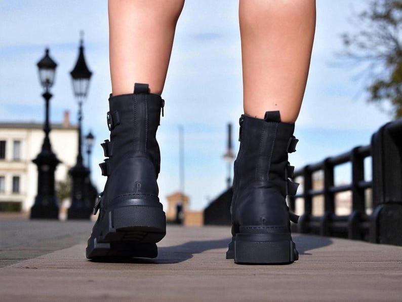 May include: Black ankle boots with multiple buckles and a chunky sole. The boots have a zipper on the side and are made of a matte material. The boots are worn on a wooden surface.