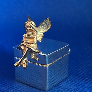 Könnte beinhalten: Eine silberne Schachtel mit einer goldenen Feenfigur darauf. Die Fee sitzt mit ausgebreiteten Flügeln. Die Schachtel hat einen Deckel mit Scharnier.