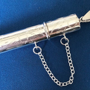 May include: A silver, engraved, cylindrical pendant with a chain attached. The pendant is hollow and has a hinged lid.