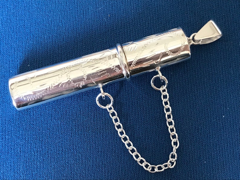 May include: A silver, engraved, cylindrical pendant with a chain attached. The pendant is hollow and has a hinged lid.