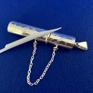May include: A silver pendant with a small, cylindrical container attached to a chain. The container has an intricate engraved design and a white stick inside.