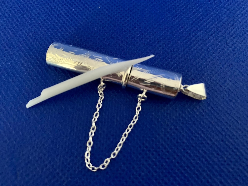 May include: A silver pendant with a small, cylindrical container attached to a chain. The container has an intricate engraved design and a white stick inside.