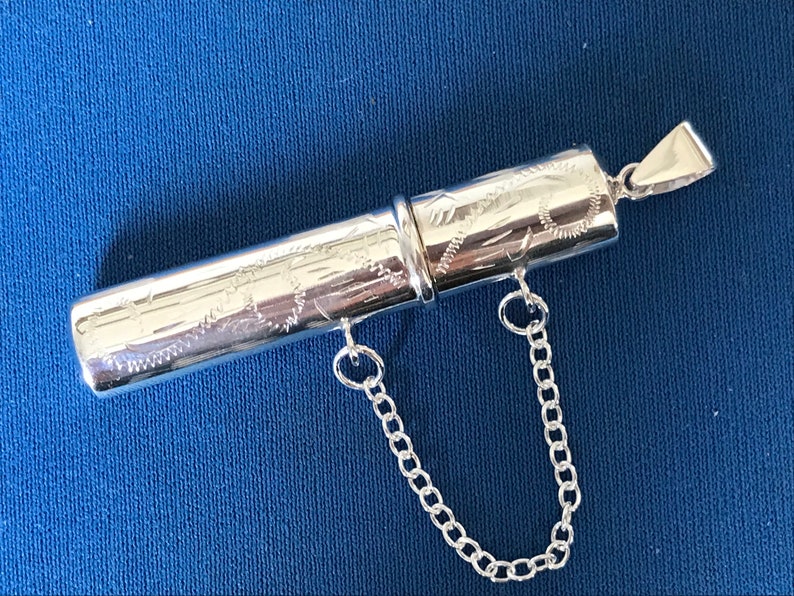 May include: A silver, engraved, cylindrical pendant with a chain attached. The pendant is made of two pieces that fit together.