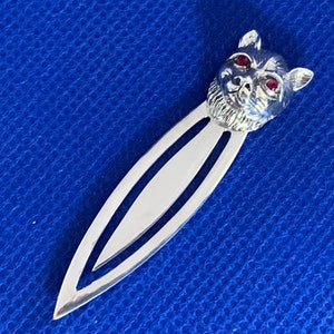 May include: A silver-colored bookmark with a cat's head design. The cat's head has red gemstone eyes and detailed fur. The bookmark is on a blue background.