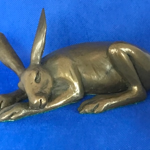 Puede incluir: Una escultura de bronce de una liebre acostada con la cabeza apoyada en las patas. La liebre está representada en un estilo realista, con pelo y rasgos detallados.