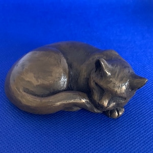 以下が含まれることがあります： リラックスしたポーズで丸まっている、眠っている猫のブロンズ色の彫刻。猫の足は体に折りたたまれ、頭はそっと足の上に置かれています。彫刻は青い背景に配置されています。