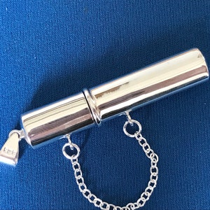 May include: A silver, cylindrical, hinged pill box with a chain attached. The box has a small inscription that reads "925".