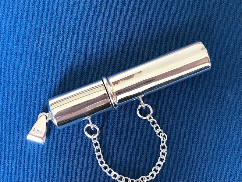 May include: A silver, cylindrical, hinged pill box with a chain attached. The box has a small inscription that reads "925".