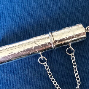 May include: A silver, engraved, cylindrical pendant with a chain attached. The pendant is made of two pieces that screw together.