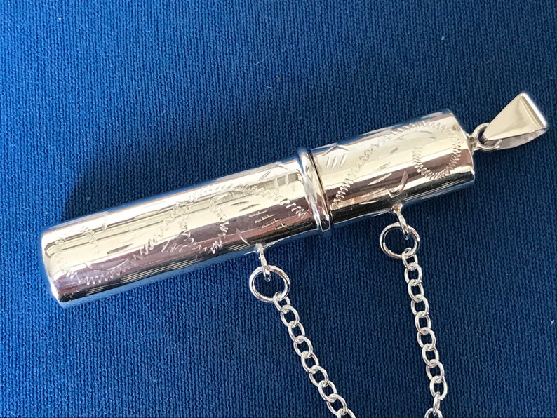 May include: A silver, engraved, cylindrical pendant with a chain attached. The pendant is made of two pieces that screw together.