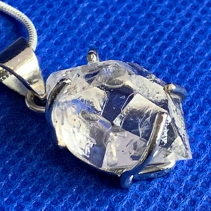 Op de afbeelding: Een heldere kristallen hanger met een zilveren setting, hangend aan een zilveren ketting. De kristal is gefacetteerd en heeft een geometrische vorm.