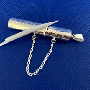 May include: A silver pendant with a small, cylindrical container attached to a chain. The container has an intricate engraved design and a small, white stick inside.
