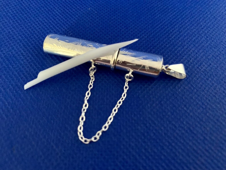 May include: A silver pendant with a small, cylindrical container attached to a chain. The container has an intricate engraved design and a small, white stick inside.