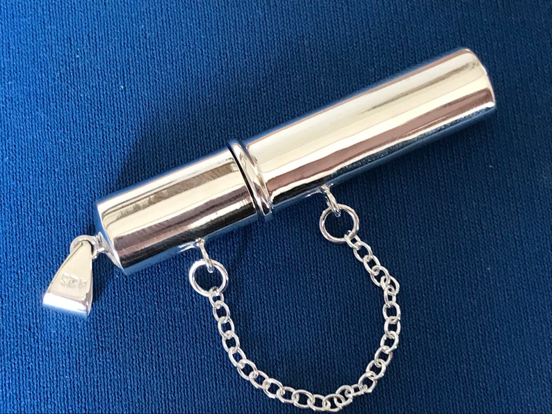 May include: A silver, cylindrical, two-piece pill box with a chain attached. The box is engraved with the number "925" on the bottom.