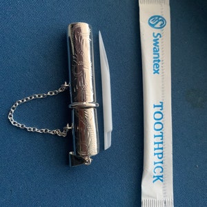 May include: A silver toothpick holder with an engraved design and a chain. The holder is next to a white toothpick wrapper with the text "Swantex Toothpick" printed in blue.