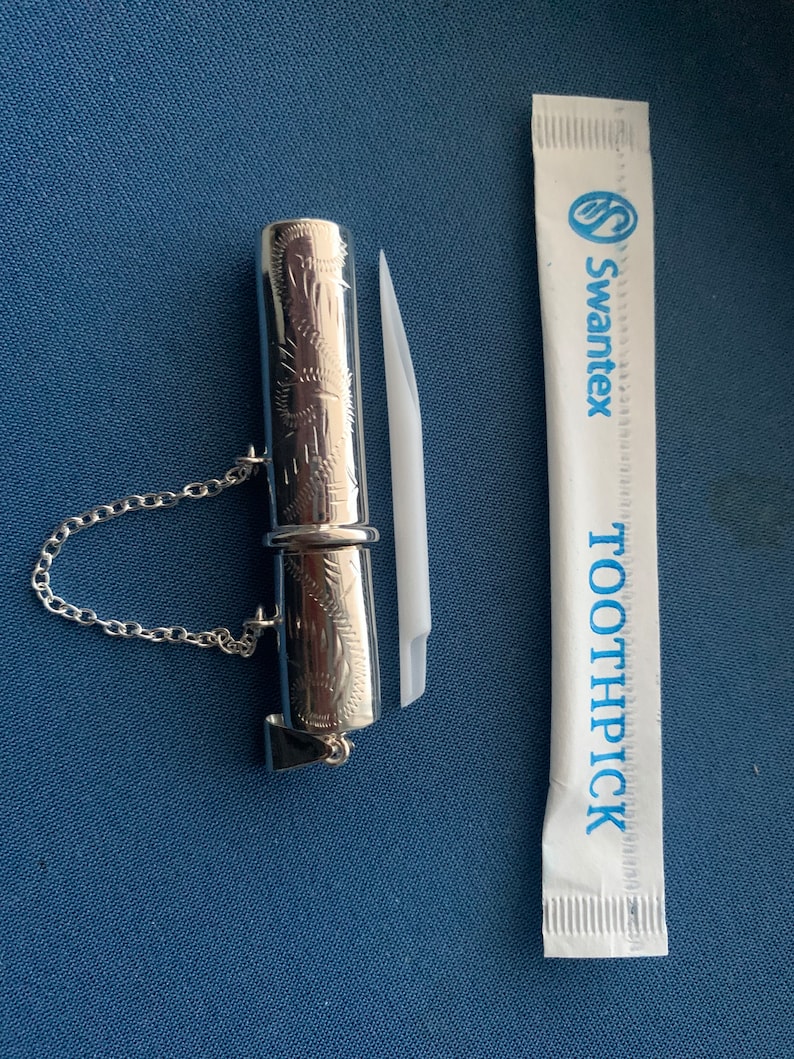 May include: A silver toothpick holder with an engraved design and a chain. The holder is next to a white toothpick wrapper with the text "Swantex Toothpick" printed in blue.