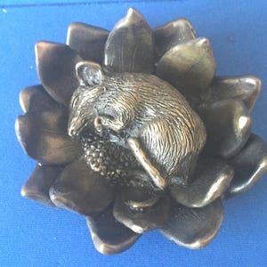 May include: A bronze-colored decorative object featuring a detailed sculpture of a mouse nestled within a stylized flower. The flower has multiple petals, and the mouse is curled up. The overall design has a vintage aesthetic.
