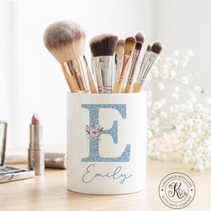 May include: White ceramic makeup brush holder with a large blue glitter letter "E" and the name "Emily" in script. The letter is decorated with a floral design. The holder contains various makeup brushes.