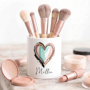 May include: A white ceramic makeup brush holder featuring a painted heart design in shades of teal, pink, and gold glitter with the name Millie printed in black script. The holder contains five makeup brushes with rose gold ferrules and tan bristles.