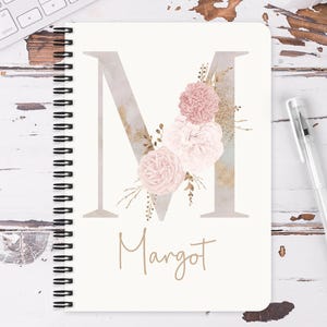 May include: A cream-coloured spiral-bound notebook with a large, watercolour-style letter "M" on the cover. It features pink floral accents and the name "Margot" below. A clear pen is placed next to the notebook.