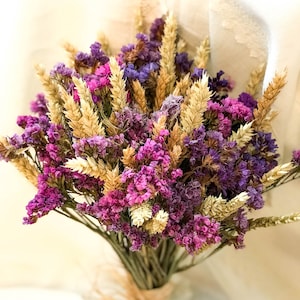 May include: A bouquet of dried flowers in shades of purple and pink, with wheat stalks woven throughout. The bouquet is tied with twine.