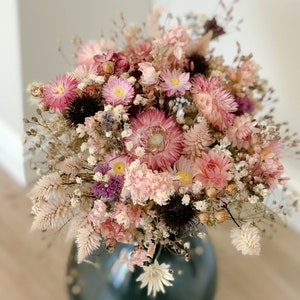 Può includere: Un bouquet di fiori secchi in tonalità rosa e bianche, disposti in un vaso blu scuro. Il bouquet comprende garofani, gipsofila e altri fiori secchi.