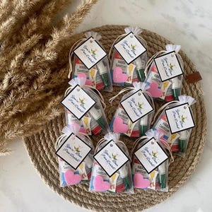 Pode incluir: Uma coleção de pequenas sacolas de presente, cada uma contendo um tubo de desinfetante para as mãos e um item em forma de coração rosa. Cada sacola tem uma etiqueta com texto e é amarrada com uma fita. As sacolas estão dispostas em um tapete circular tecido.