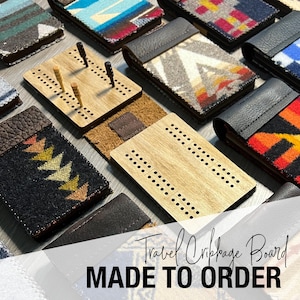 May include: A collection of travel cribbage boards with various patterned fabric covers and wooden game boards. The boards are displayed with pegs inserted, showcasing the game's design. The text "Travel Cribbage Board MADE TO ORDER" is visible.