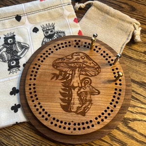 May include: A wooden cribbage board with a mushroom design. The board has a brown stain and is decorated with a mushroom and a smaller mushroom growing from the ground. The board has a circular shape and is surrounded by a ring of holes.
