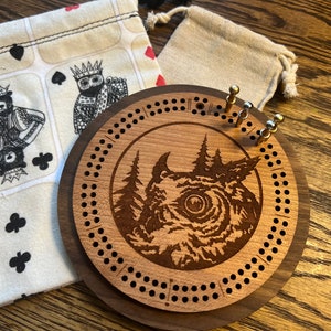 May include: A wooden cribbage board with an owl design and a dark brown wood frame. The board has a circular design with holes for pegs and a central image of an owl in a forest setting.