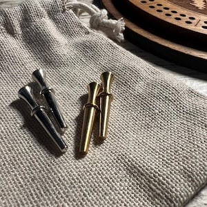 May include: A set of cribbage pegs in silver and gold tones, resting on a beige linen bag. The pegs have a classic design with a flared top and a slender body. A wooden cribbage board is visible in the background.