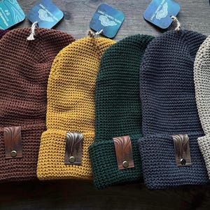 May include: Six knitted beanies in different colors, including burgundy, brown, yellow, green, blue, and beige. Each beanie has a leather patch with a design on the side.
