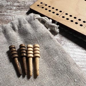 May include: A wooden cribbage board with drilled holes and a set of wooden pegs in a burlap bag. The pegs are various shades of brown and tan. The board and bag are on a light-colored wooden surface.